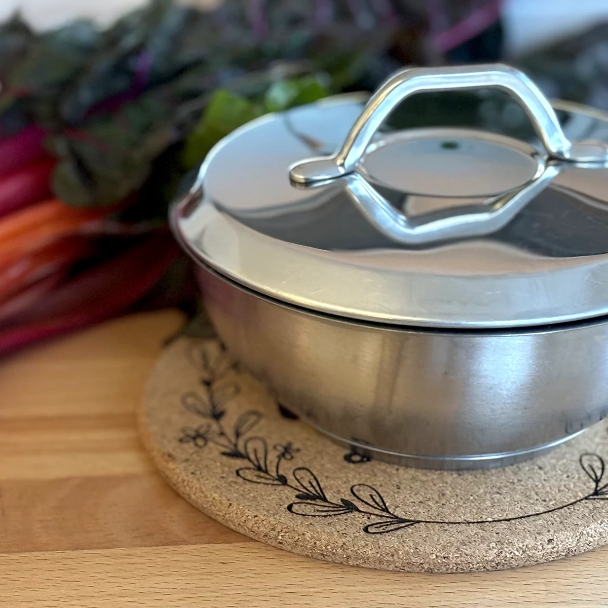 Kochtopf mit Deckel auf rundem Korkuntersetzer mit graviertem floralem Motiv.