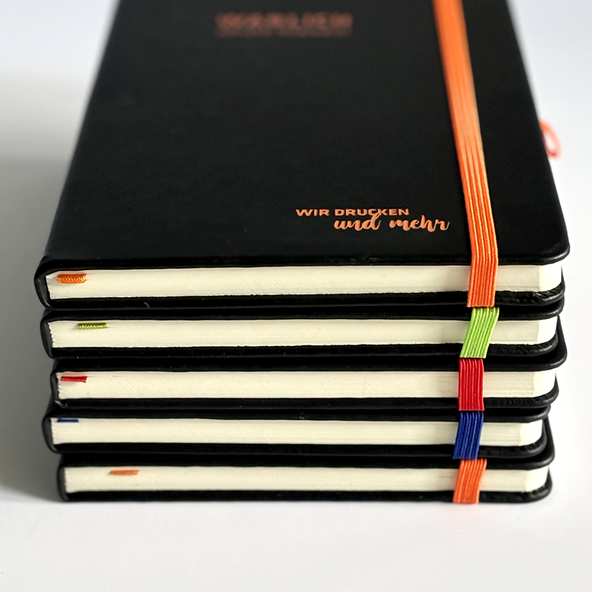 Gestapelte schwarze Notizbücher mit graviertem Logo und farbigen Gummibändern in Orange, Grün, Rot und Blau, seitlich fotografiert, auf hellem Hintergrund.