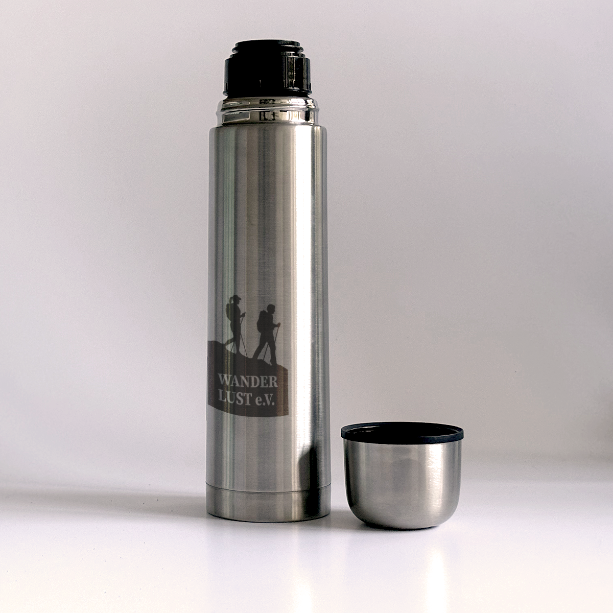 Edelstahl-Thermoskanne mit graviertem Wandermotiv und dem Schriftzug „WANDERLUST e.V.“, daneben abgenommener Deckel, der als Becher dient.