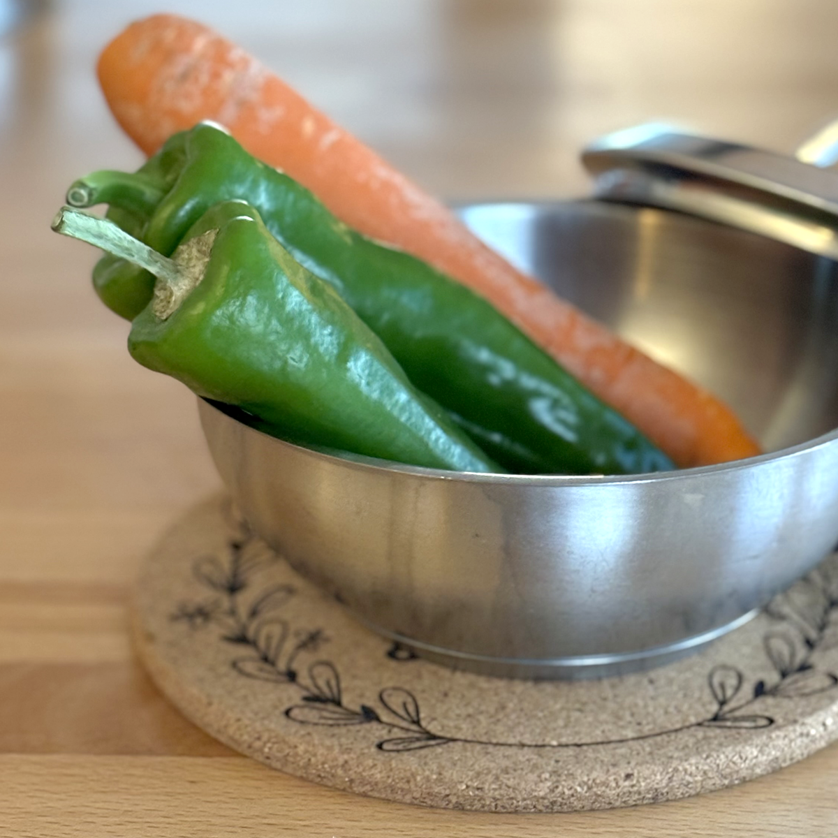 Kochtopf ohne Deckel mit Gemüse auf rundem Korkuntersetzer mit graviertem floralem Motiv.