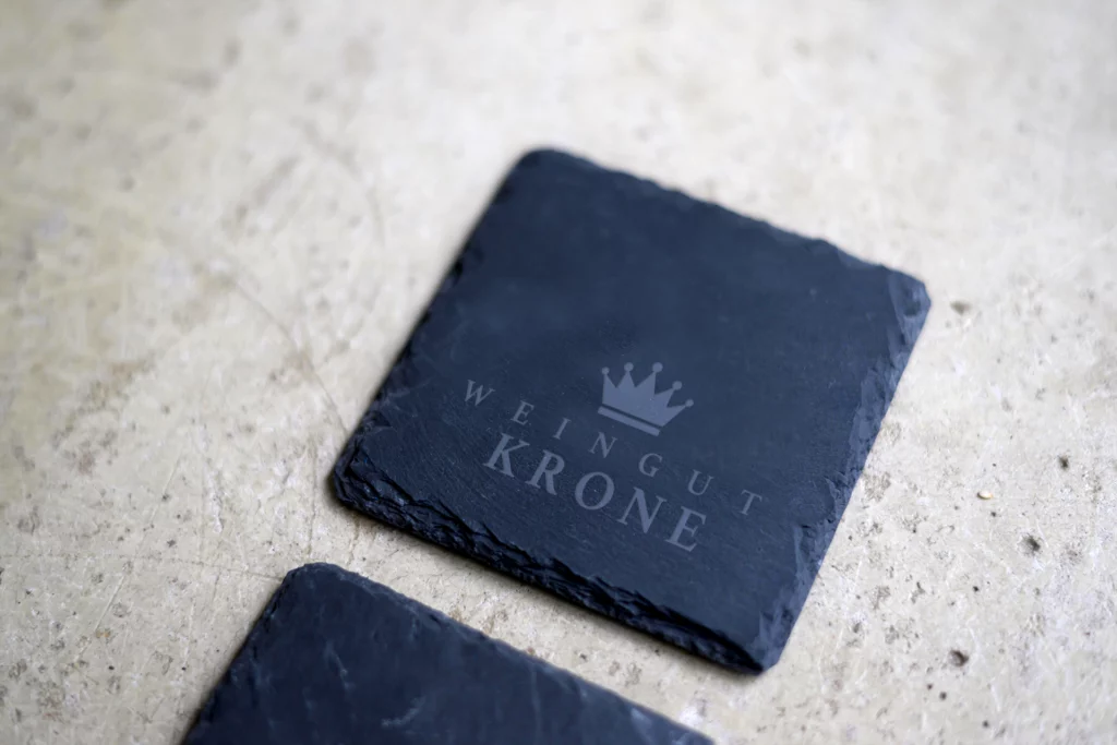 Quadratischer Schieferuntersetzer mit graviertem Logo „Weingut Krone“ und einer Krone darüber, auf heller Steinoberfläche liegend.