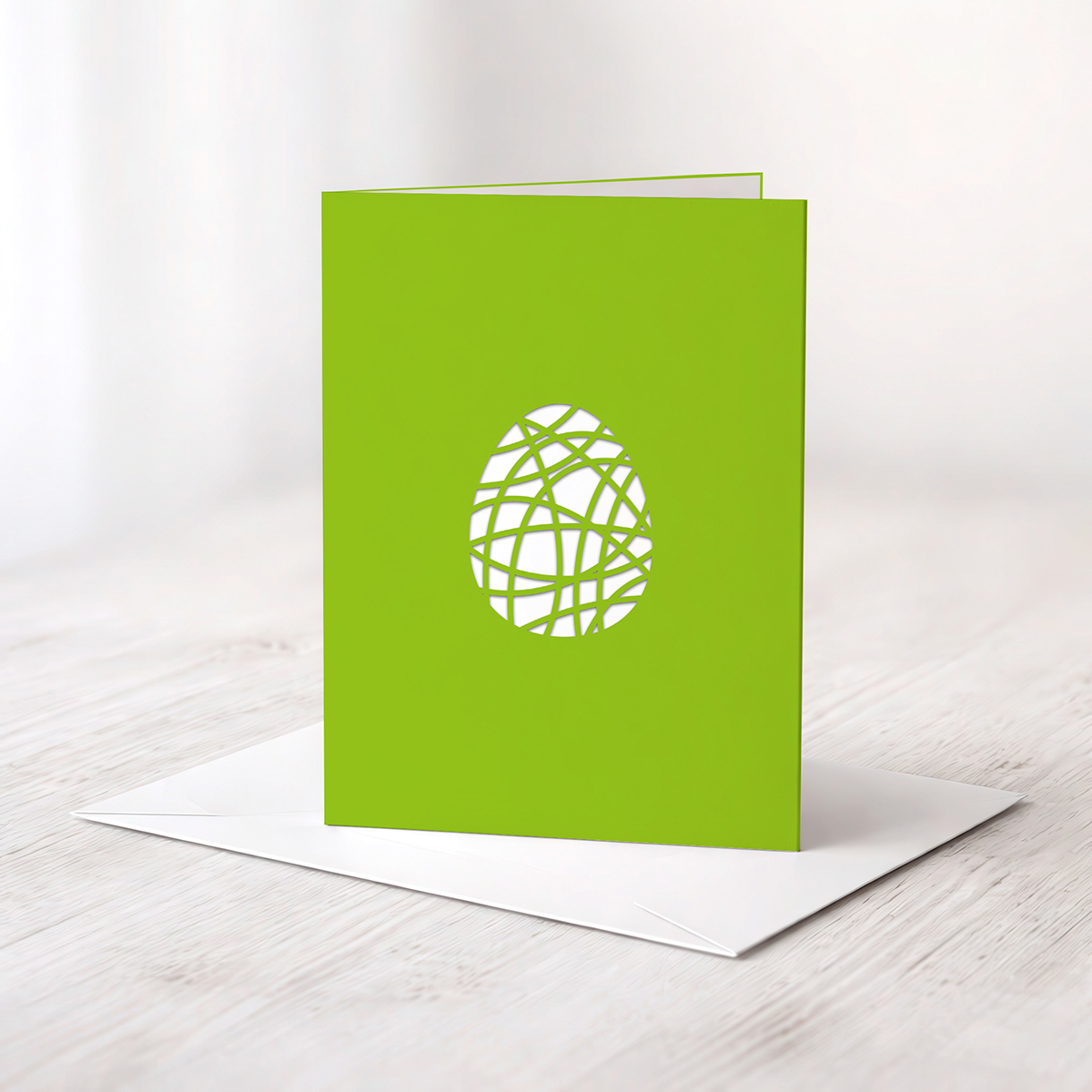 Eine grüne Karte mit einem ausgelaserten Ei aus Ornamenten liegt auf einem weißen Umschlag.